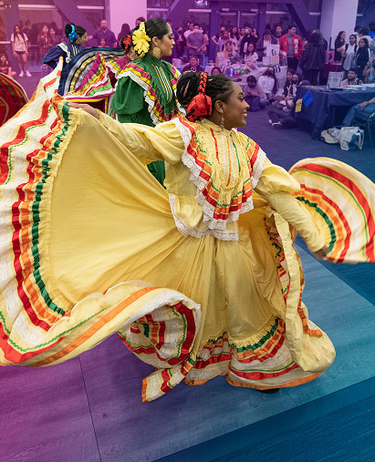 Woman are performing a dance in front of an audience. In the front of the picture is a woman holding up an end of her yellow dress.